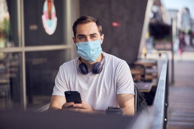 Genç maskeli bir adam boş terasta kahvenin tadını çıkarıyor. Elinde bir fincan kahve var.