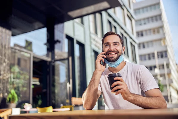 Man with a lowered mask is drinking coffee and talking on the smartphone
