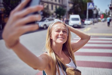 Diş teli takan kızıl saçlı neşeli kız güneşten gözlerini kısarak selfie çekiyor.  