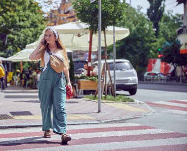 Güzel, güler yüzlü, mutlu, çilli kız yaya geçidinde yürürken akıllı telefonla konuşuyor ve uzak bir yere bakıyor.  