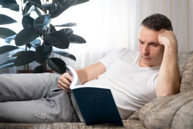 Boş zaman, eğitim ve ana konsept: Gülümseyen bir adam kitap okur ve evinde kanepede oturur. Dinlen, rahatla..