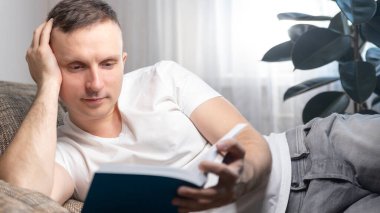 Boş zaman, eğitim ve ana konsept: Gülümseyen bir adam kitap okur ve evinde kanepede oturur. Dinlen, rahatla..