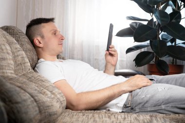 Genç, gülümseyen bir adam evdeki akıllı telefondan sosyal ağları kontrol ediyor. Gündelik giysiler içindeki şık çocuk, boşluğun kopyası.