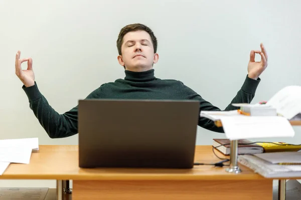 Relaxed corporate man sitting with closed eyes contemplating at work desk with laptop in office. Guy relaxing, relieves stress, gets positive thinking with yoga exercise. Stress free workplace concept