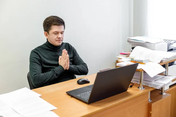 Relaxed corporate man sitting with closed eyes contemplating at work desk with laptop in office. Guy relaxing, relieves stress, gets positive thinking with yoga exercise. Stress free workplace concept