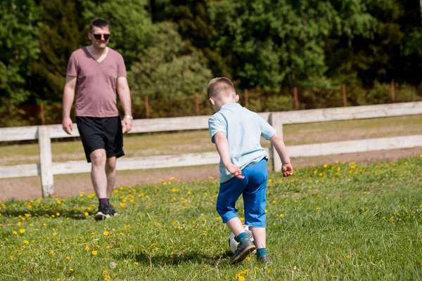 英俊的爸爸与他的小可爱的太阳在绿色草草坪上玩美式足球 男孩用蓝色衬衣踢足球 — 图库照片