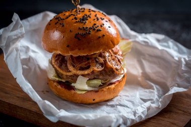Büyük bir duble burger, soğan ve peynir, karanlık bir arka plan üzerinde bir sürü. Ahşap Tepsi ve kağıt.