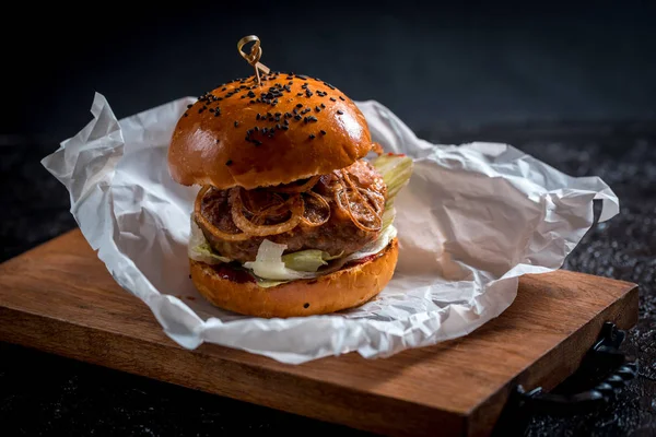 A large double burger, a lot of onions and cheese, on a dark background. wooden tray and paper.