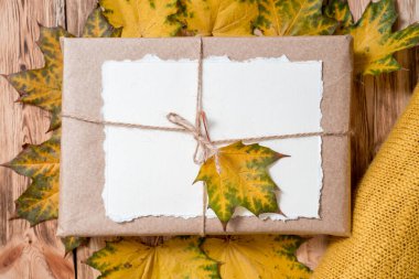 Kraft ambalajlı hediye kutusu. Notların için yırtılmış kağıt. Sonbahar parlak kuru yapraklar. Ahşap arka planda sarı örülmüş süveter. Sıcak sonbahar konsepti. Uzayı kopyala düz yat.