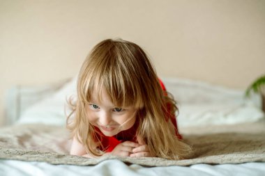 Yatağa oturmuş gülümseyen bebek. Parlak bir yatak odasında sevimli kızıl saçlı kız oynuyor. Komik ve şakacı. Kırmızı elbiseli. Mutlu çocukluk.