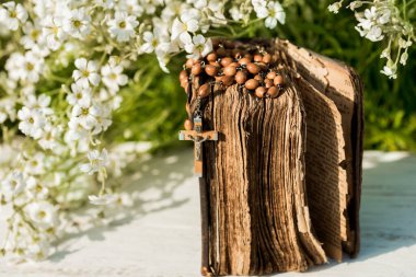 İncil 'in başında dua ederken ellerim katlandı. Ahşap zemin. El ve tespih, dua, sarı sayfalı eski kitap. Arka planda beyaz çiçekler. Bahçede. Dua ve din kavramı.