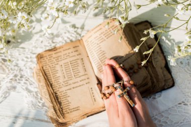 İncil 'in başında dua ederken ellerim katlandı. Ahşap zemin. El ve tespih, dua, sarı sayfalı eski kitap. Arka planda beyaz çiçekler. Bahçede. Dua ve din kavramı.