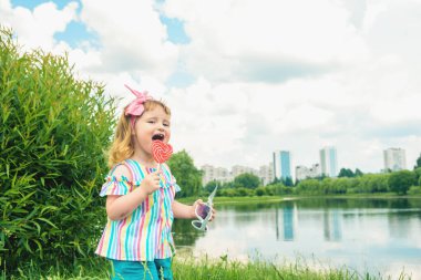 Modaya uygun bir kız bebek, güneş gözlüğü, kalp gibi lolipop. Üç yaşında küçük bir kız, yaz günü parkta yürürken renkli bir girdap şekerini yalıyor..