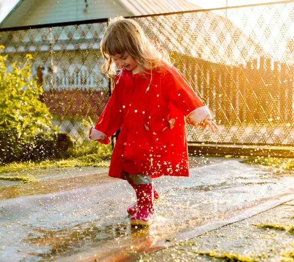 Cute girl in a red jacket is jumping in the puddle.The setting warm summer or autumn sun. summer in the village