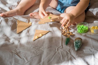 Bir çocuk kartondan bir Noel ağacı oyuyor. Noel süsü, el yapımı ve sıfır atık. Çocuk evdeki yatakta el işi yapıyor. Div. en üstteki görünüm