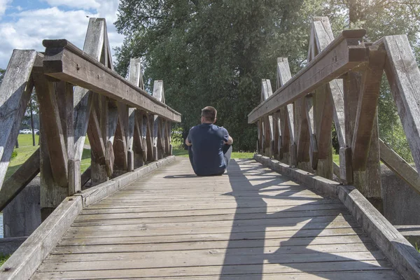 The man sits with his back turned on an old wooden bridge