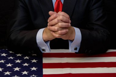 Politikacı-işadamı, Amerikan bayrağıyla kaplı masada otururken parmaklarını kilit altına aldı.