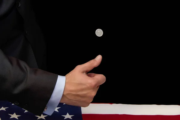 A man in a suit tosses up a 5-cent coin with his hand on a dark background, the coffin is covered with a flag, concept: the price of a penny