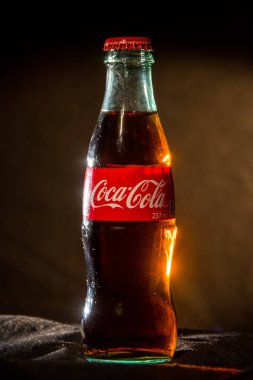 Coca-Cola yazıtlı metal kapak, koyu bir arka plan, renk parlaması ve parlama üzerinde bir cam şişe önünde havaya düşüyor; Beyaz Rusya, Minsk, 26 Temmuz 2019.