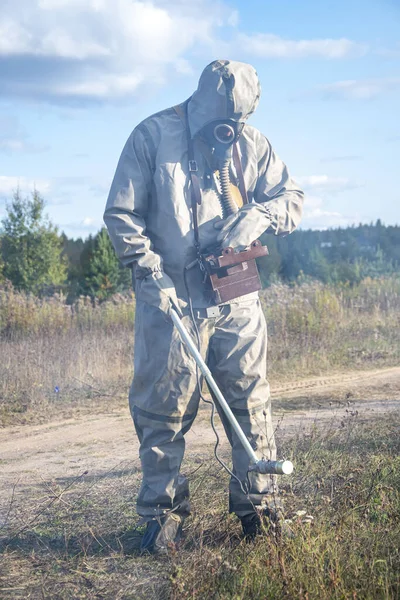 Ein Soldat in einem Chemikalienschutzanzug überprüft das radioaktive Gebiet mit einem Dosimeter vor dem Hintergrund wilder Vegetation und des Himmels mit Wolken. Rauch schädlicher Dämpfe breitet sich aus. — Stockbild Ein Soldat Einem Chemikalienschutzanzug Überprüft Das Radioaktive Gebiet Mit Einem — Stockfoto