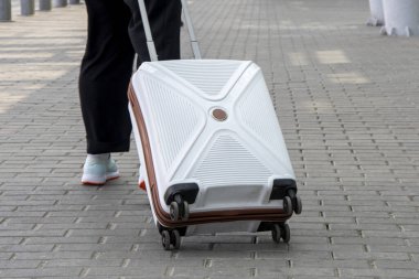 Bir kadın tekerlekli beyaz bir bavulu yuvarlıyor, yakın plan. Konsept: Seyahatlerde ve seyahatlerde el bagajı, yolda eşyalar, tatile çıkmak..