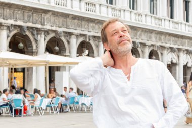 A man aged 50-55 in a white shirt stands in a square in Venice, enjoying the day