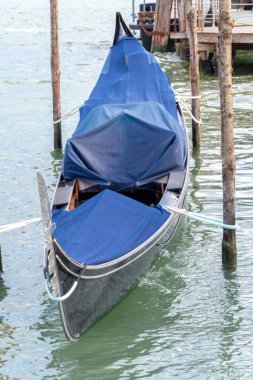A classic Venetian gondola with a blue cover is moored to wooden piles in the water