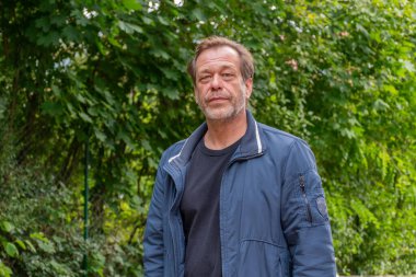 Portrait of an elderly man aged 50-55 wearing a blue jacket against a backdrop of green trees