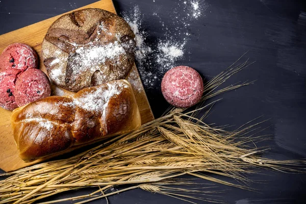 Sortiment čerstvě upečených chlebů s ušima z pšenice a čokoládově červených sušenek na tmavém dřevěném stole. Horní pohled. — Stock fotografie