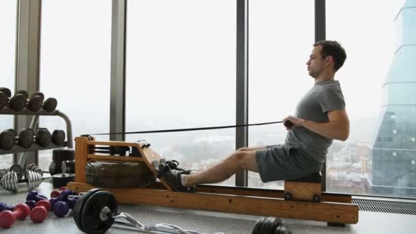 A young fit man trains on a rowing machine in a gym - view from the side — Stock Video