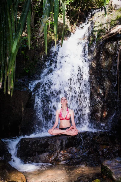 Mujer joven practicando Yoga meditando bajo Cascada —  Fotos de Stock