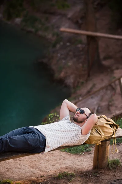 徒步旅行者时尚男子躺在湖边的木凳上 — 图库照片