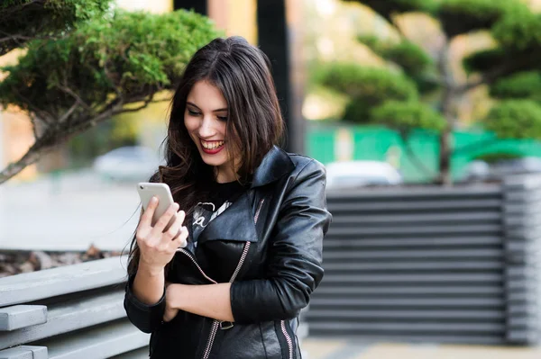 Hermosa joven morena está enviando mensajes de texto en su teléfono inteligente mientras está sentada en el banco en el parque de la ciudad — Foto de Stock