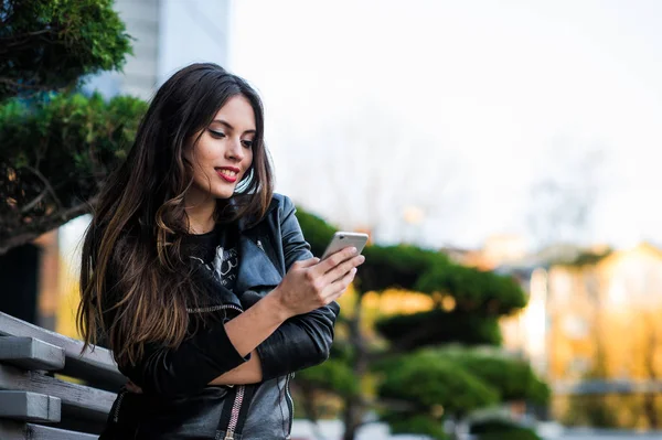 Chica caminando y mensajes de texto en el teléfono inteligente en la calle con una chaqueta de cuero en verano — Foto de Stock