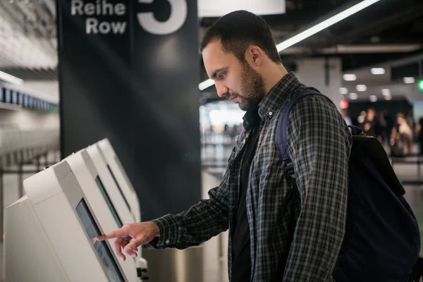 Junger Mann mit Rucksack berührt interaktives Display mit Selbstbedienungsautomat, macht Self-Check-in für Flug oder kauft Flugtickets am Automaten im modernen Flughafenterminal — Stockfoto