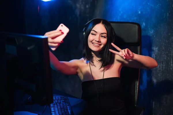 Alegre gamer streamer menina vestindo fone de ouvido e sentado perto de seu computador pc tomando selfie no telefone durante o jogo online — Fotografia de Stock