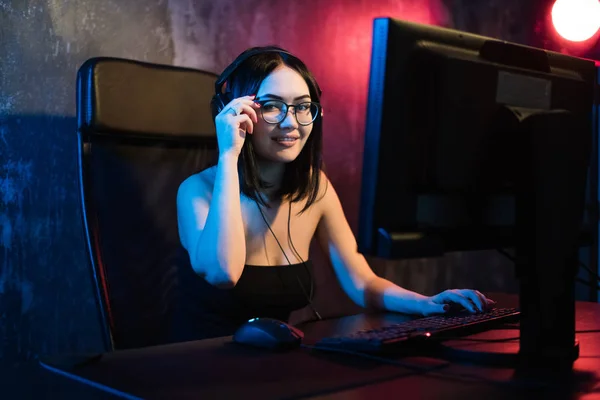 Beautiful young girl wearing glasses and gaming headset plays online game on gaming PC in dark area. Streaming online games on internet