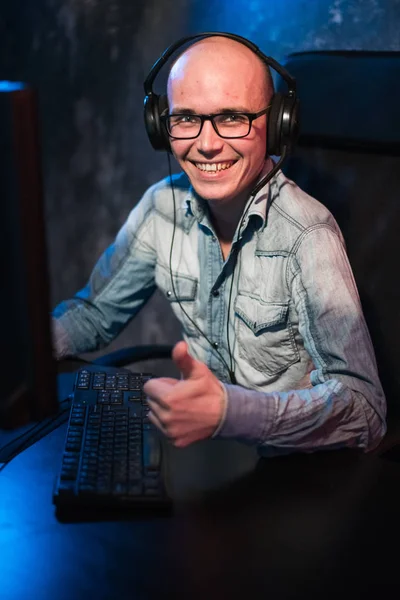 Retrato de un joven jugador alegre jugando videojuegos en casa en una computadora. Gamer en los auriculares es feliz y mostrando los pulgares hacia arriba gesto porque él ha ganado el juego. Juegos de ordenador Concepto . —  Fotos de Stock