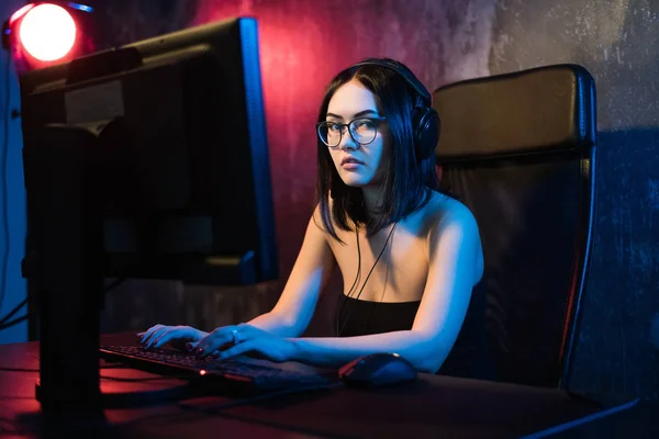 Hermosa chica joven con gafas y auriculares de juego juega juego en línea en PC de juego en el área oscura. Streaming juegos en línea en Internet —  Fotos de Stock
