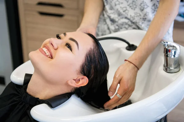 Young brunette girl smiling and enjoying hair washing in hairdressing salon. — Stock Photo, Image