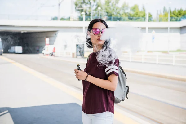Elegante chica fumando un cigarrillo electrónico mientras camina por la ciudad. — Foto de Stock