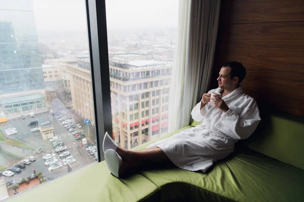 Joven hombre guapo y despreocupado cerca de la ventana moderna de longitud completa disfrutando de una taza de café mientras mira hacia el exterior, buenos días, primer día en una nueva ciudad, libre de trabajo y deber, viajero. Copiar espacio —  Fotos de Stock