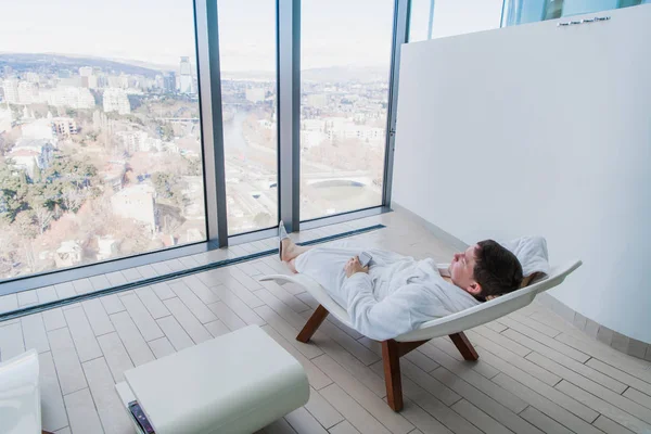 Joven descansando en una moderna tumbona frente a una gran ventana con vista a la ciudad en un moderno spa —  Fotos de Stock