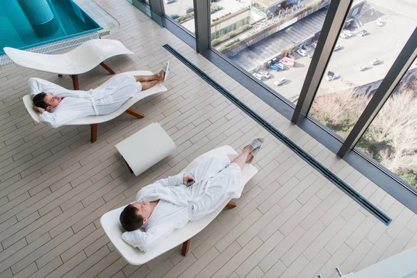 Twee jonge man dragen badjas, liggend op een ligstoel in spa salon en tegenover een groot raam met uitzicht op een grote stad met elkaar praten. Spa concept — Stockfoto