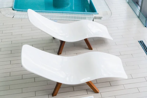 Empty two white lounge chairs inside of tiled room near swimming pool. Nobody in spa room. Deck chair for clients relaxation after spa procedures, bath house and swimming in pool. Modern and