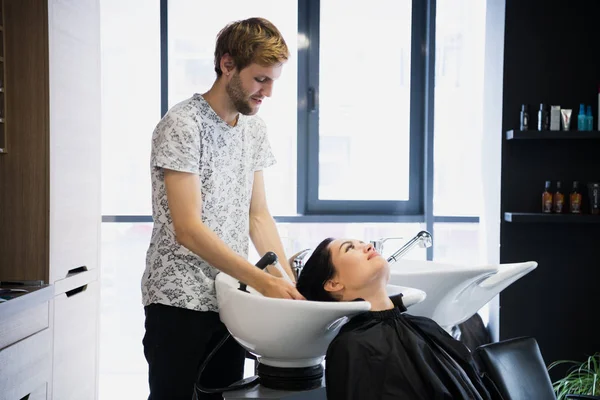 Genç hippi erkek kuaför salon bir güzel esmer kadın kadın istemcisinin saç yıkama. — Stok fotoğraf