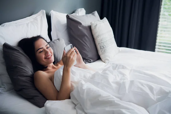 Feliz atractiva joven europea sonriendo ampliamente mientras lee el mensaje de texto de su novio usando el teléfono móvil en la cama, sin usar nada, cubriendo el cuerpo con una manta — Foto de Stock