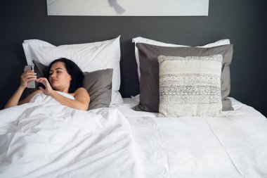 Esmer kız yatakta sevgilisi mobil, süre üzerinde metin mesajı okuyor. Battaniye ile örtülü neşeli genç kadın önce cep telefonuyla video mesajı sonra uyku izliyor.