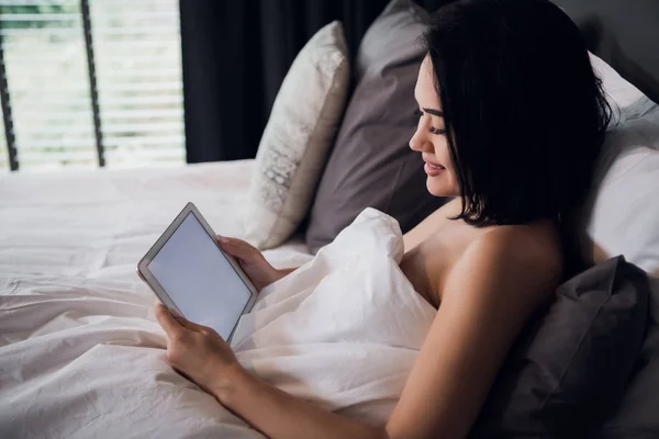 Atractiva chica morena sosteniendo tableta digital con pantalla en blanco y sonriendo en el dormitorio — Foto de Stock