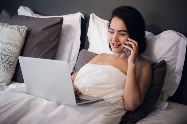 Vista lateral de la joven sonriente con ordenador portátil y teléfono en la cama — Foto de Stock